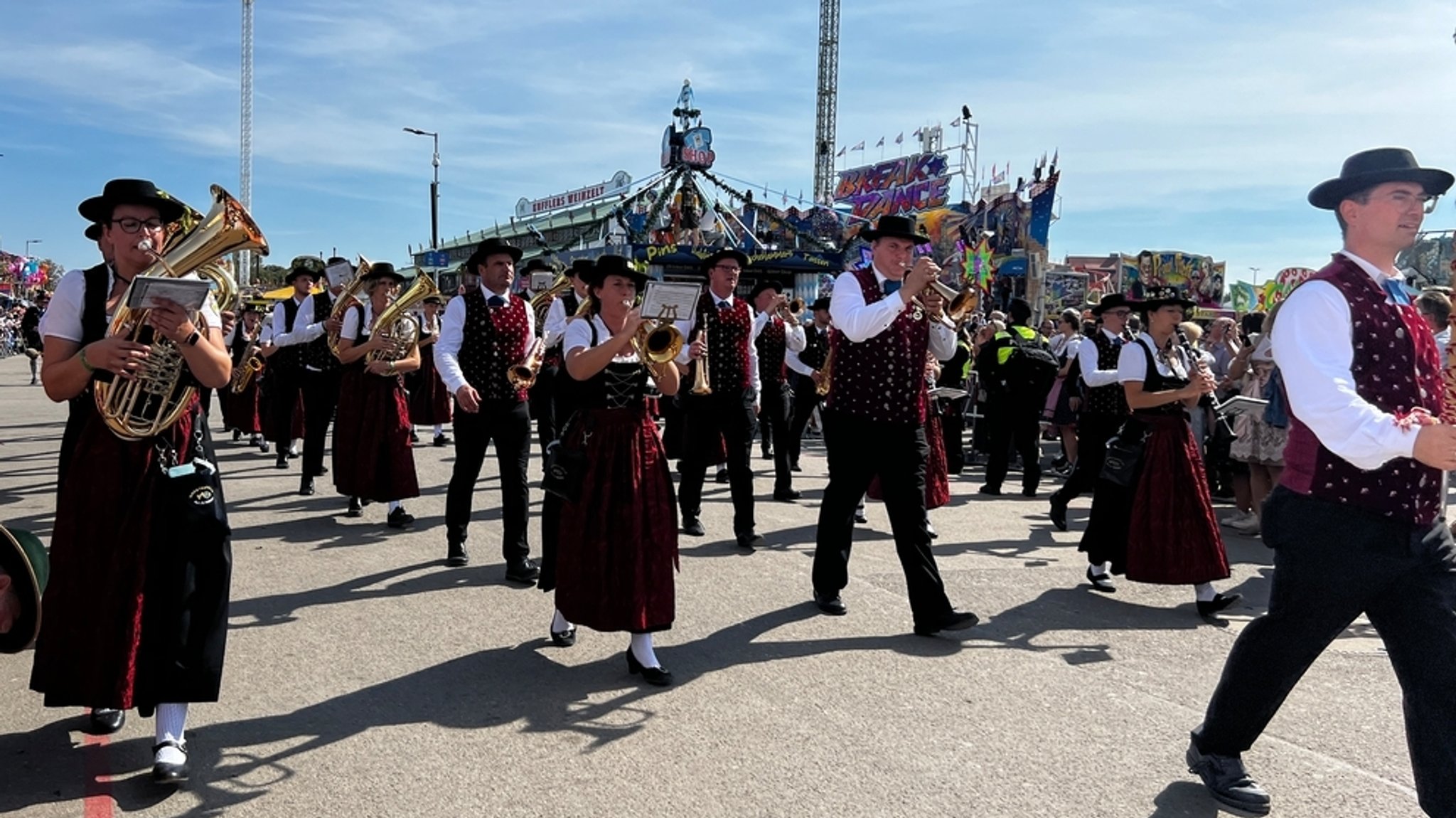 Eine Blasmusikkapelle in Tracht läuft mit dem Trachten- und Schützenzug auf der Theresienwiese ein. Im Hintergrund Buden und Fahrgeschäfte des Oktoberfests