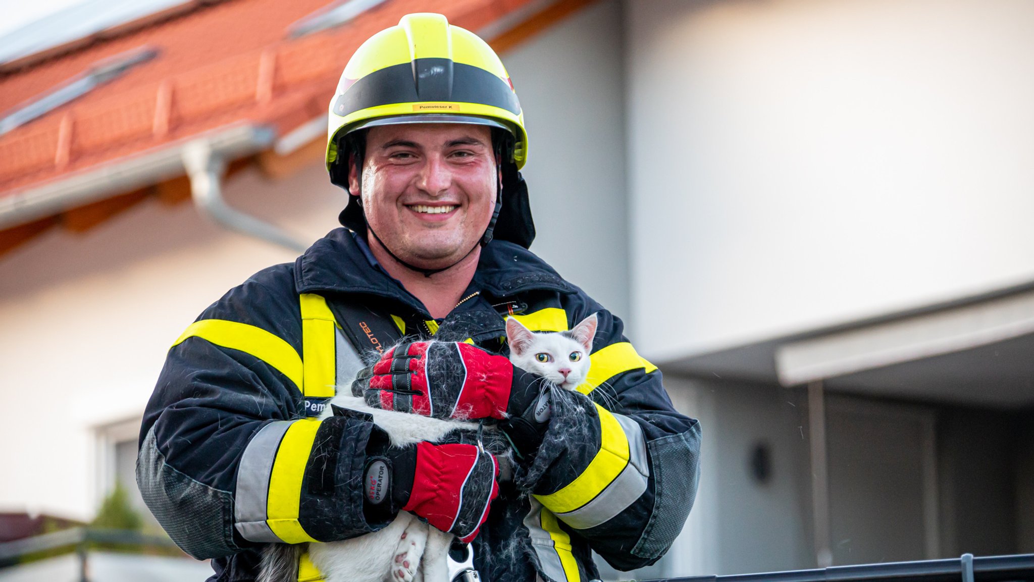 Feuerwehrmann Kevin Pemwieser in voller Montur mit Helm und Schutzanzug. Er hält Kater Oleg in den Armen, eine weiße Kurzhaar-Katze mit einem grauen Fleck zwischen den spitzen Ohren