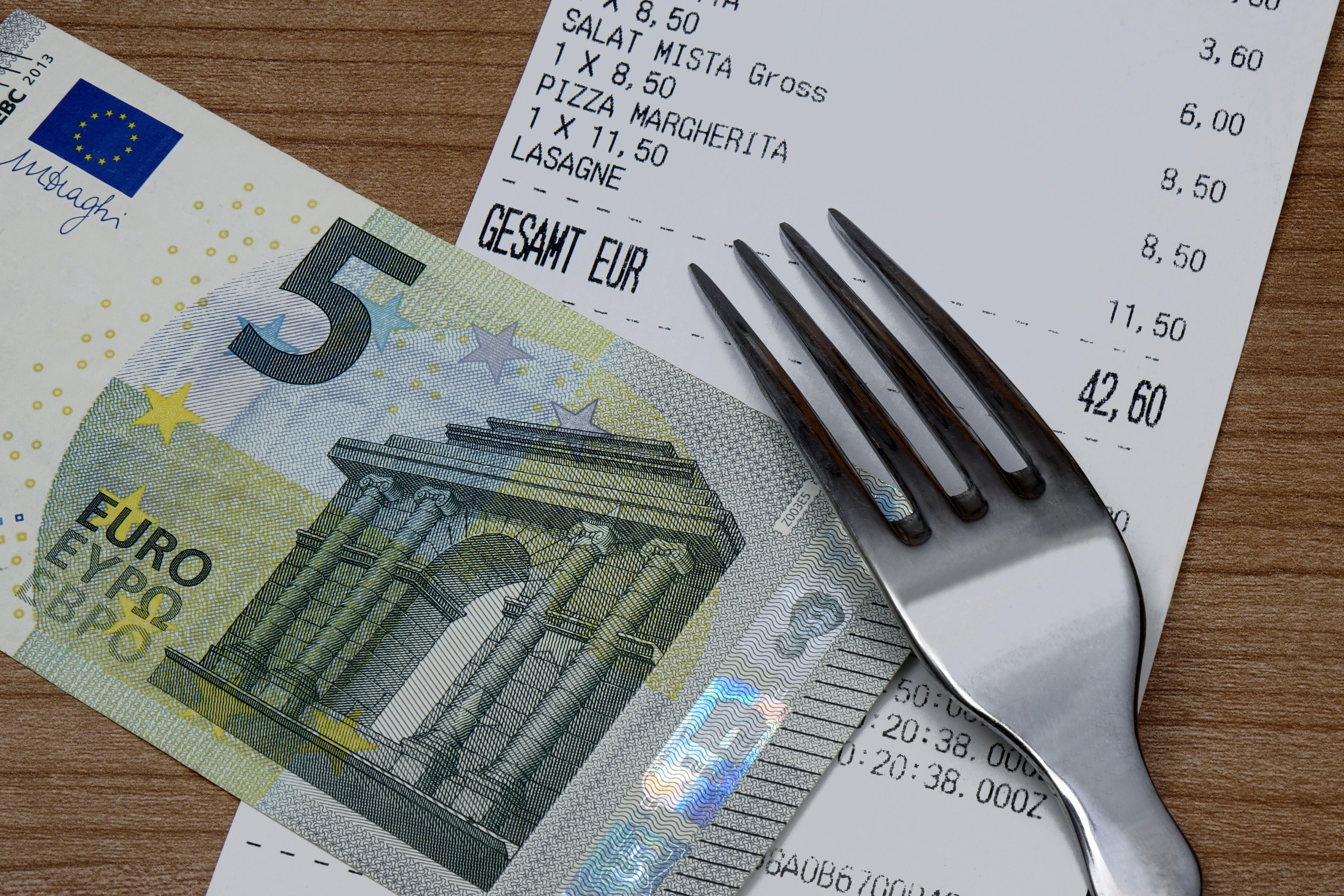 Symbolbild: Ein Gaststätten-Kassenzettel, auf dem ein Fünf-Euro-Schein und ein Gabel liegen.