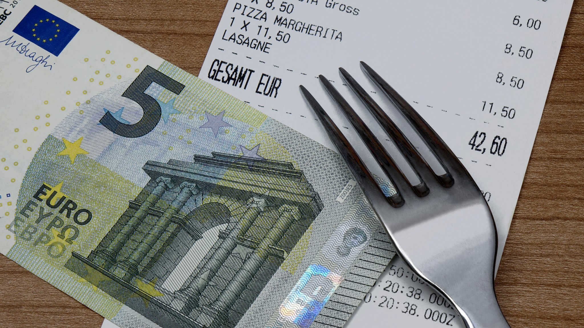 Symbolbild: Ein Gaststätten-Kassenzettel, auf dem ein Fünf-Euro-Schein und ein Gabel liegen.