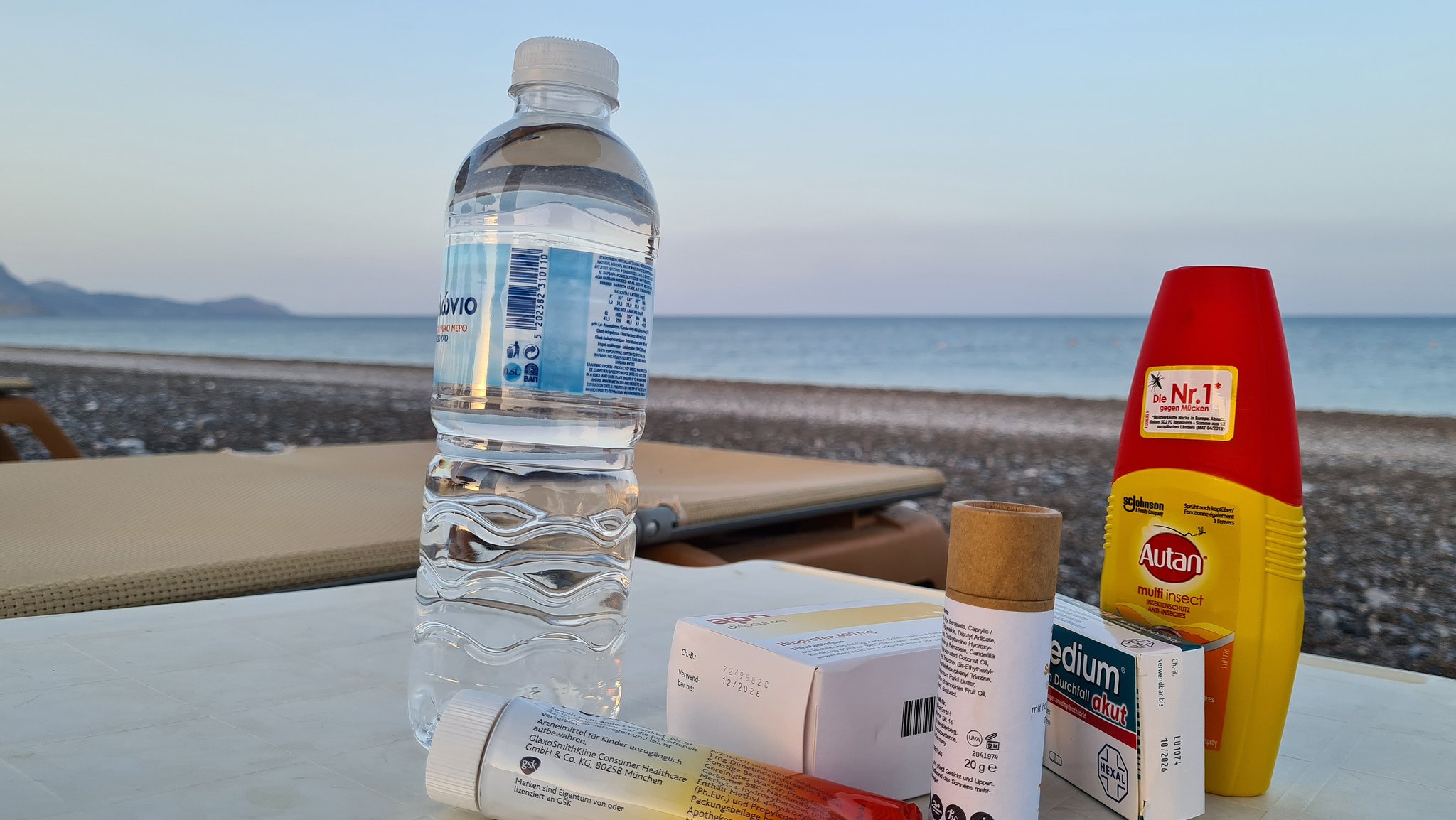 Auf einem Tisch am Strand liegen eine Flasche Wasser, Arzneimittel wie Durchfalltabletten und Kopfschmerztabletten und ein Insektenschutzmittel.