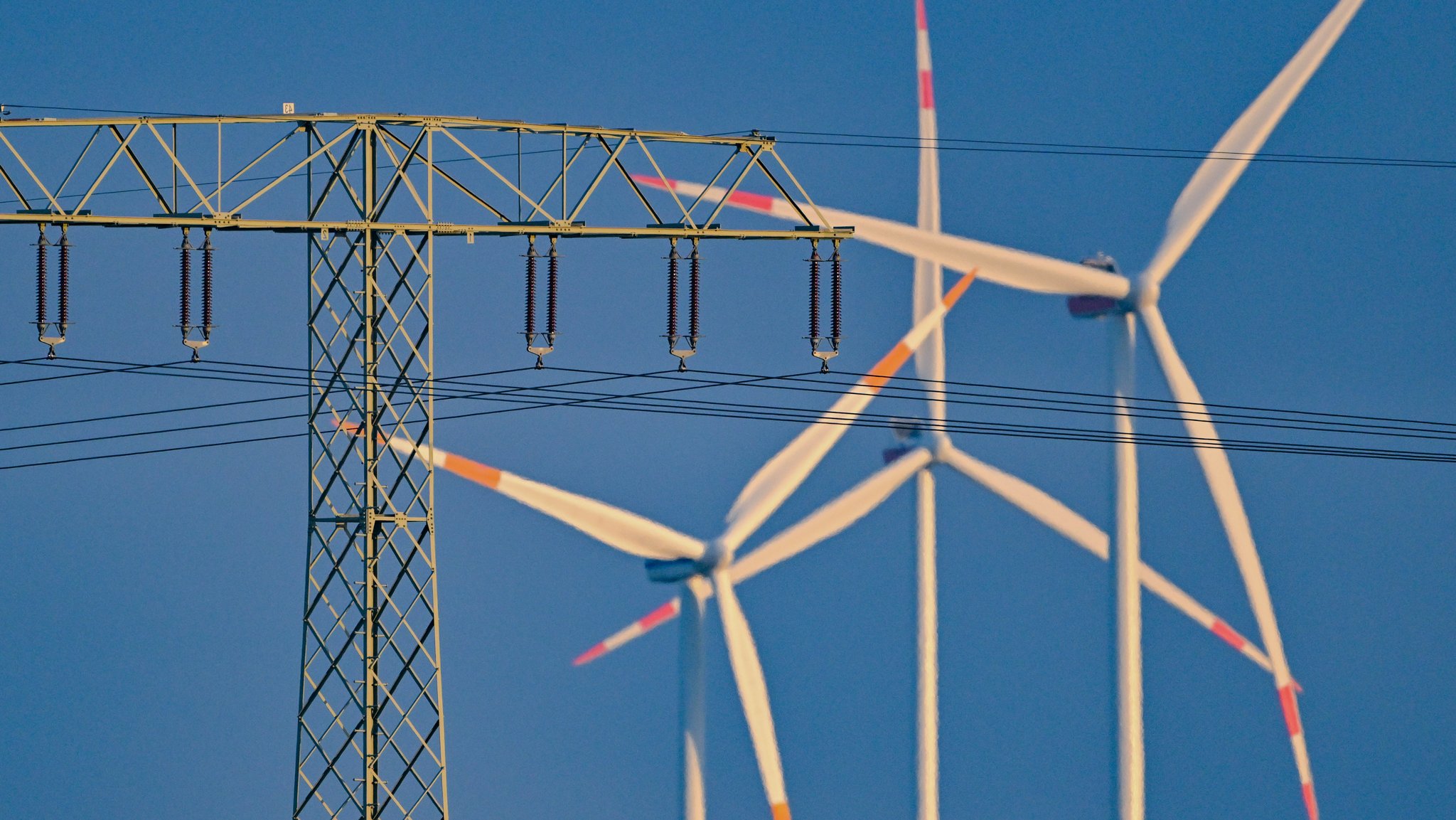 Ein Strommast mit einer Hochspannungsleitung steht vor Windenergieanlagen