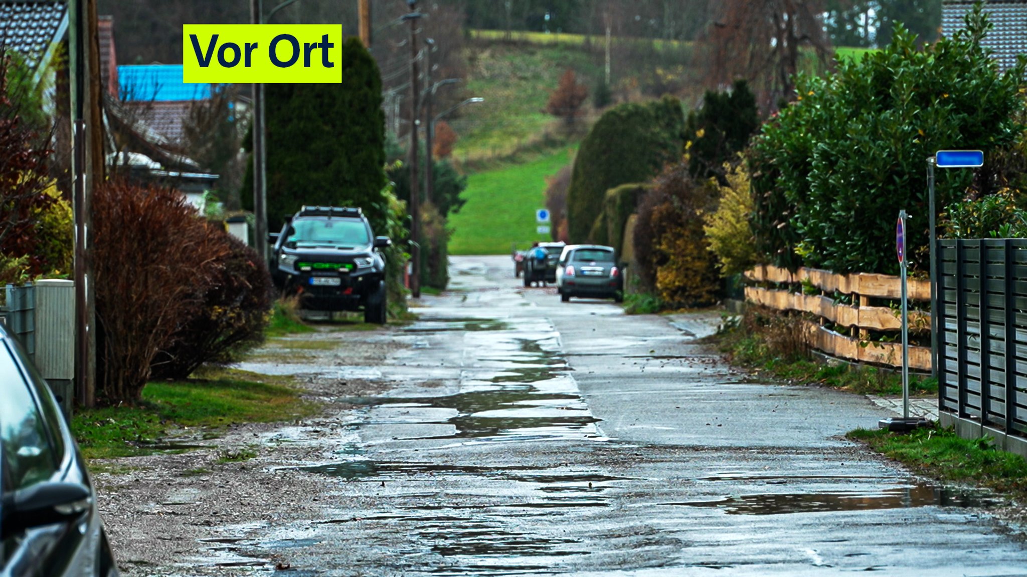 Eine asphaltierte Straße in Eichenau mit vereinzelten Straßenlaternen und Kies und Gras am Straßenrand, in Unebenheiten sammelt sich das Wasser.