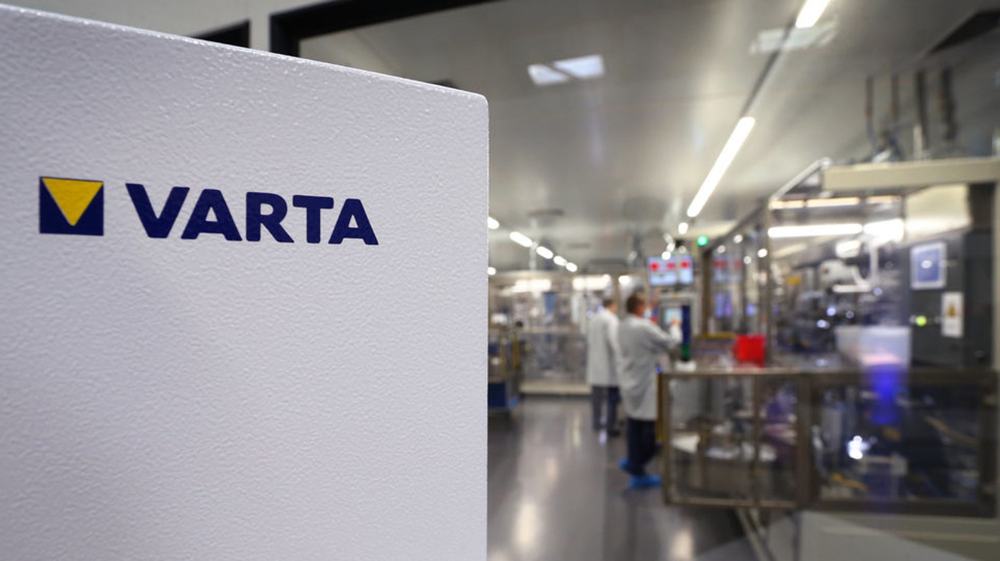 Das Firmenlogo des Batterieherstellers VARTA, im Hintergrund ein Produktionsraum mit Mitarbeitern