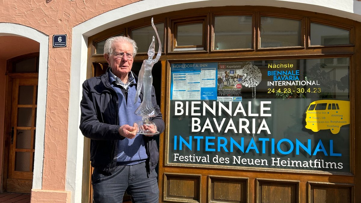 Der Altbürgermeister Günther Knoblauch steht vor dem Bürgerbüro mit einer gläsernen Figur in den Händen, an der Fassade wird die Biennale Bavaria beworben