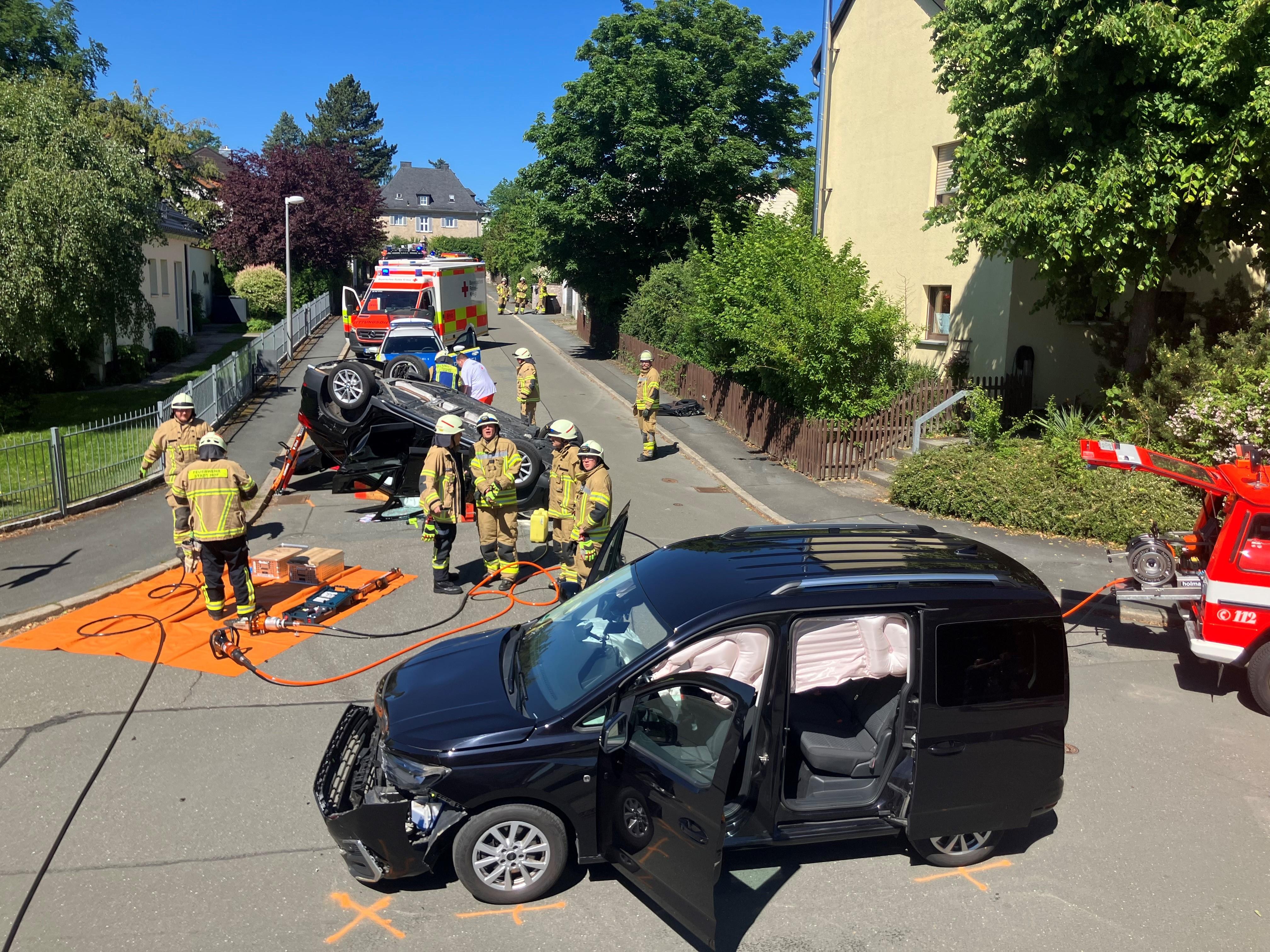 Auf einer Kreuzung liegt ein Auto auf dem Dach, ein anderes ist im Frontbereich beschädigt, dazwischen stehen Feuerwehrmänner.
