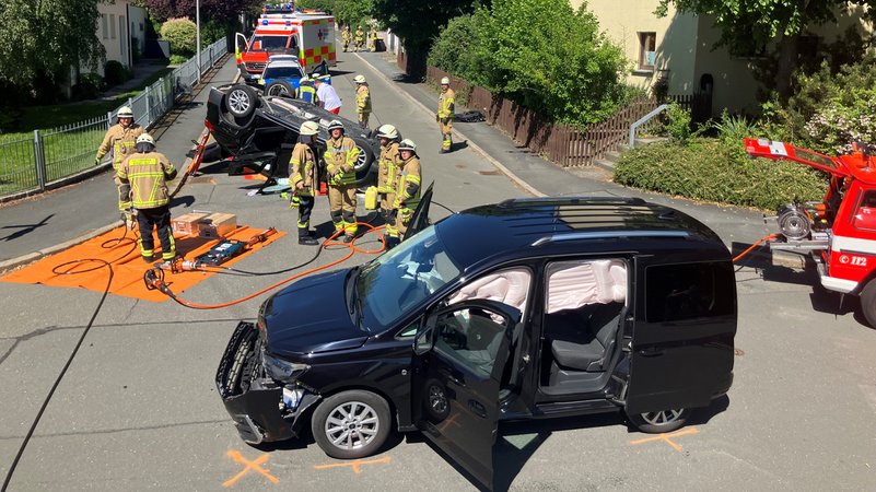 Auf einer Kreuzung liegt ein Auto auf dem Dach, ein anderes ist im Frontbereich beschädigt, dazwischen stehen Feuerwehrmänner. | Bild: Polizeiinspektion Hof Auf einer Kreuzung liegt ein Auto auf dem Dach, ein anderes ist im Frontbereich beschädigt, dazwischen stehen Feuerwehrmänner.