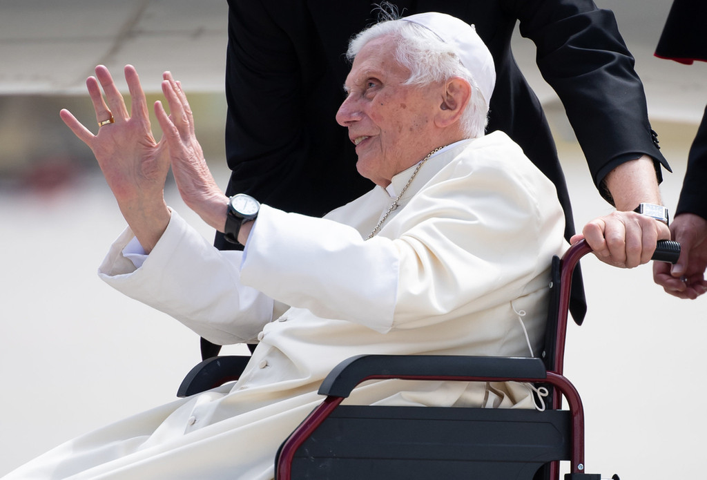 Papst Benedikt XVI. im Profil in weißem Gewand, im Rollstuhl sitzend, beide Hände zum Gruß erhoben. Archivbild aus dem Jahr 2020.