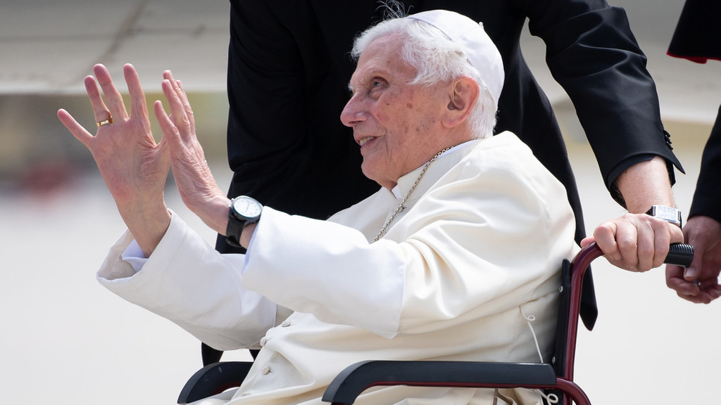 Papst Benedikt XVI. im Profil in weißem Gewand, im Rollstuhl sitzend, beide Hände zum Gruß erhoben. Archivbild aus dem Jahr 2020.