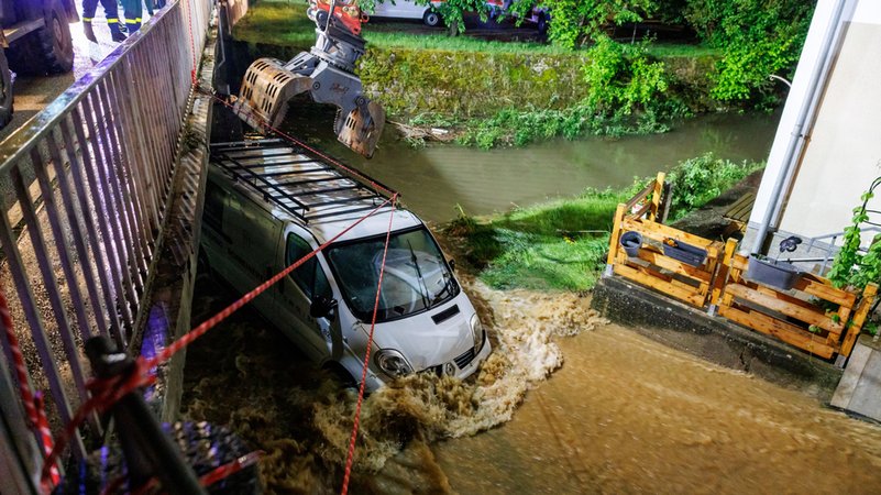 21.05.2024, Bayern, Kastl: Feuerwehrleute und ein Bagger arbeiten daran, einen Kastenwagen aus den Überflutungen vor einer Brücke zu bergen. 