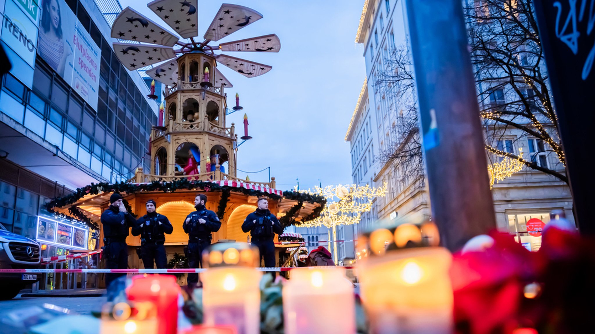 Polizisten stehen nach der Attacke auf den Magdeburger Weihnachtsmarkt vor der Weihnachtspyramide. Im Vordergrund in Unschärfe Grablichter. Am Freitag ist ein Mann mit einem Auto in Magdeburg in einen Weihnachtsmarkt gerast. Über den mutmaßlichen Attentäter gibt es erste Erkenntnisse.