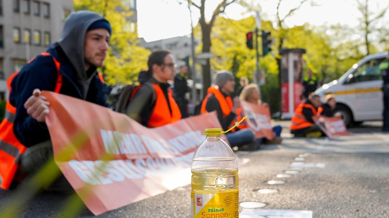 Aktivisten der Gruppe Letzte Generation bei einer Blockade-Aktion. Im Bild: Aktivisten, teils festgeklebt am Asphalt mit Bannern. Das Sonnenblumenöl zum Lösen des Klebers steht schon bereit.