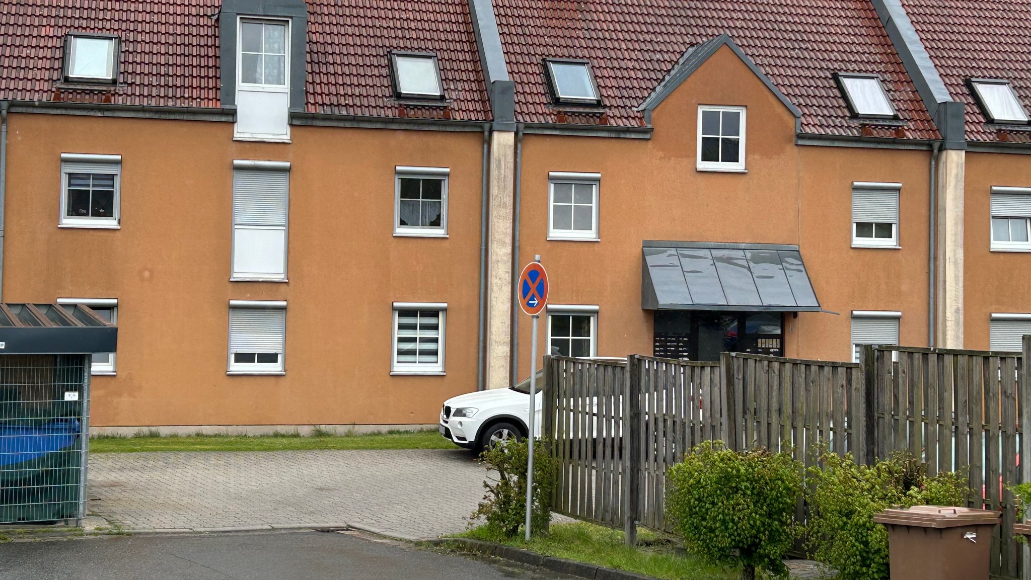 Ein braunes Mehrfamilienhaus. Im Vordergrund steht ein weißes Auto. Am Rand des Parkplatzes ist eine Biotonne und das Verkehrszeichen "absolutes Halteverbot" zu sehen.