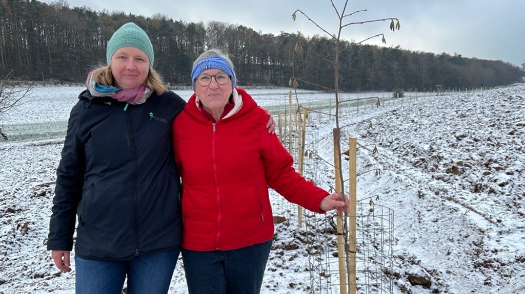 Zwei Frauen in Winterjacken stehen auf einem verschneiten Feld. Auf dem Feld sind kleine Bäume in einer Reihe gepflanzt. 