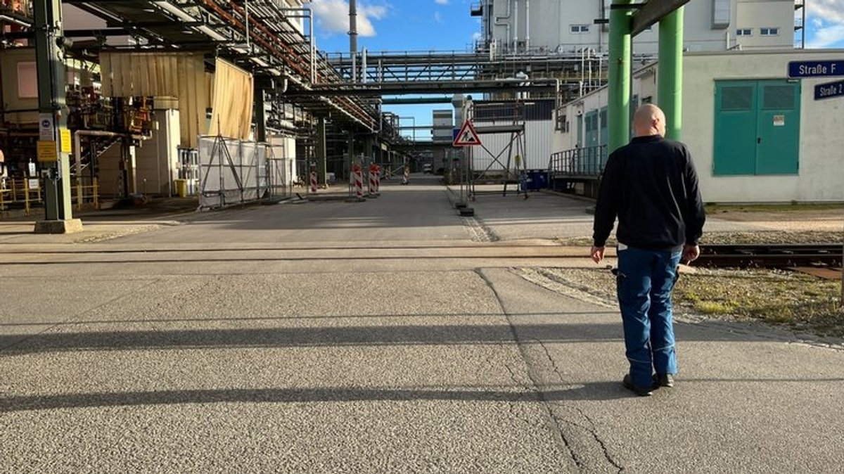 Ein 38-jähriger Mann in Arbeitshose läuft über das Gelände des Chemieparks Gendorf. Es sind viele Rohre und einige Fabrikgebäude zu sehen.