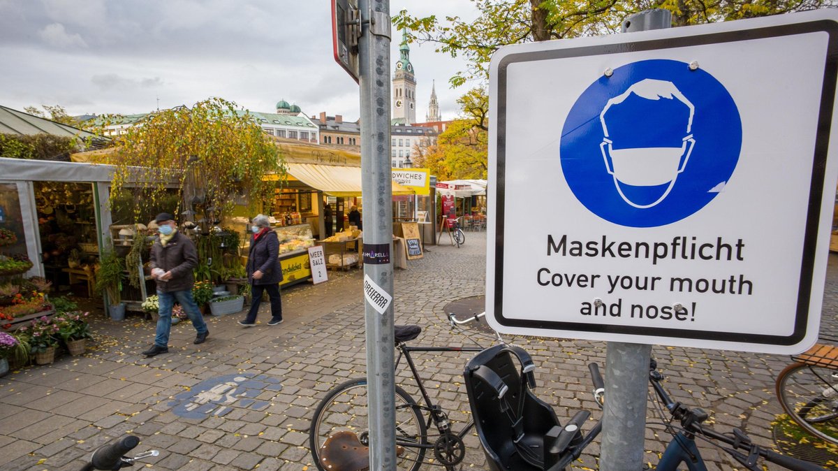 Ein Hinweisschild mit der Aufschrift "Maskenpflicht - Cover your mouth and nose!" steht am Eingang des Viktualienmarkt in der Innenstadt.