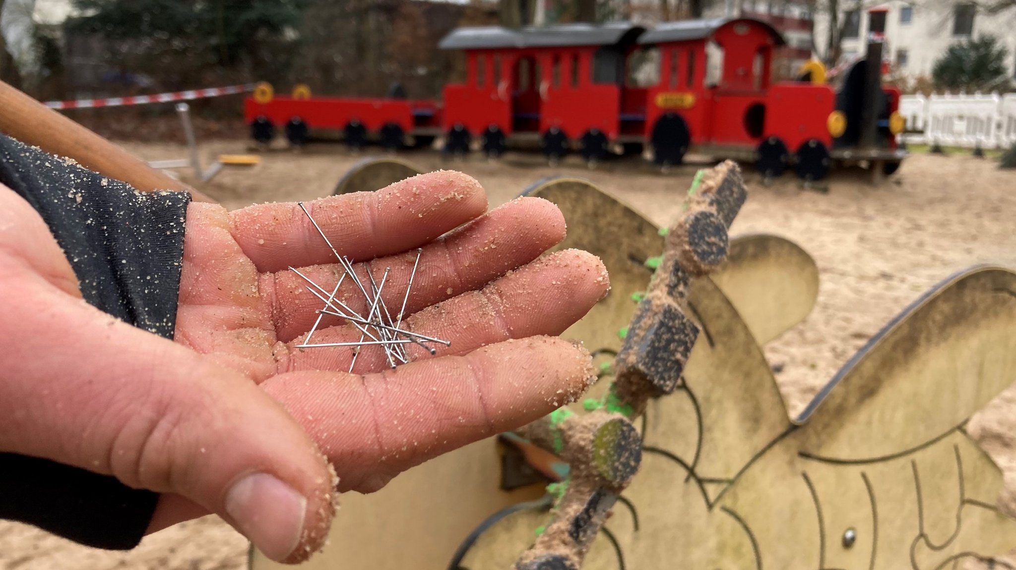 Mehrere Stecknadeln auf einer sandigen Hand, im Hintergrund eine Eisenbahn auf einem Spielplatz.