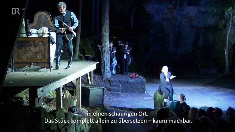 Ein Schauspieler steht auf der Bühne mit einem Gewehr in der Hand (links), während eine Frau vor der Bühne sitzt und das Stück in Gebärdensprache übersetzt