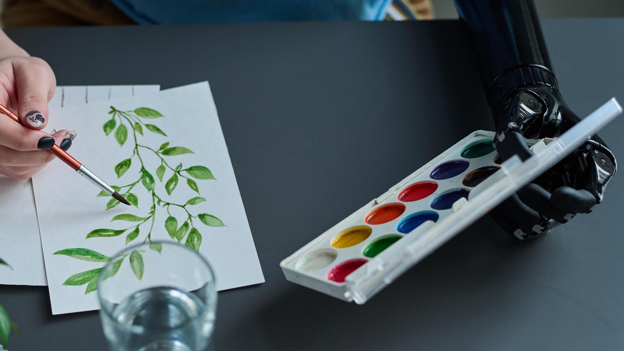 Eine junge Frau malt mit einer Hand an einem Aquarell-Bild, mit der anderen, einer bionischen Prothese, hält sie den Malkasten. (Symbolbild)
