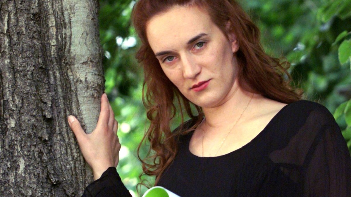 Terézia Mora steht neben einem Baum, hält ein aufgeschlagenes Büchlein in der Hand und schaut ernst in die Kamera