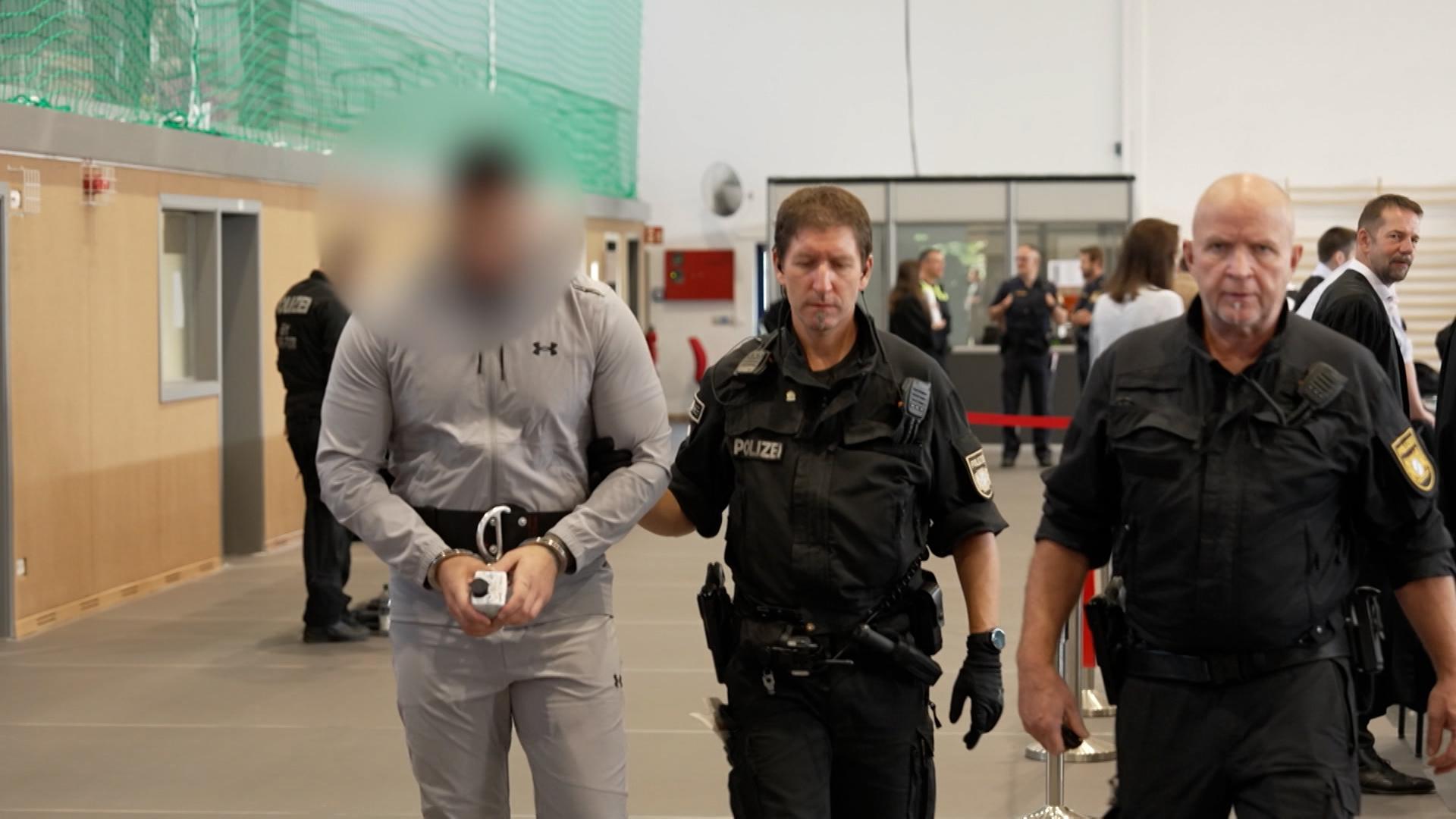 Polizisten führen einen mit Handschellen gefesselten Angeklagten zu seinem Platz in einer Turnhalle, die in einen Gerichtssaal umfunktioniert wurde.