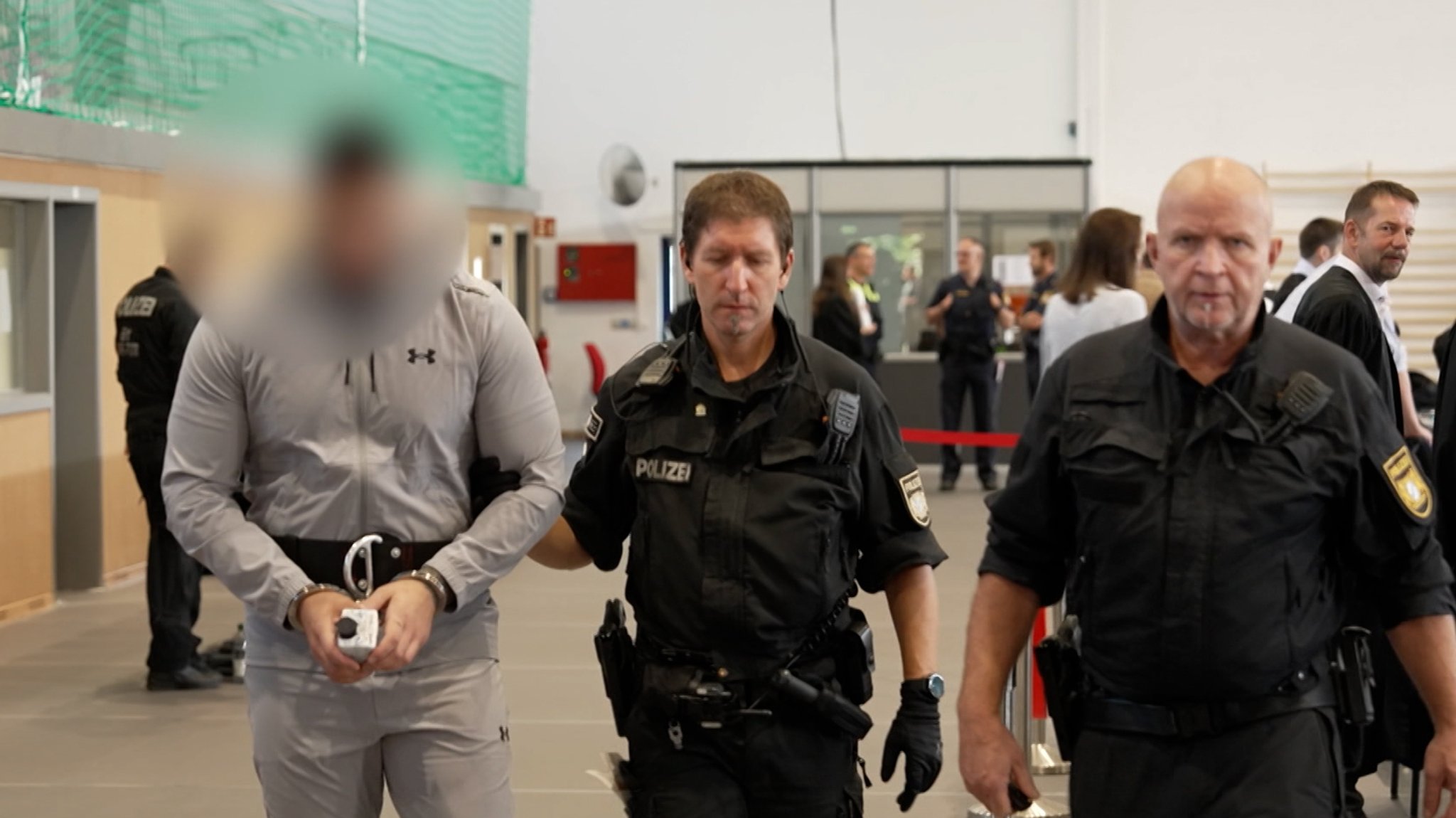 Polizisten führen einen mit Handschellen gefesselten Angeklagten zu seinem Platz in einer Turnhalle, die in einen Gerichtssaal umfunktioniert wurde.