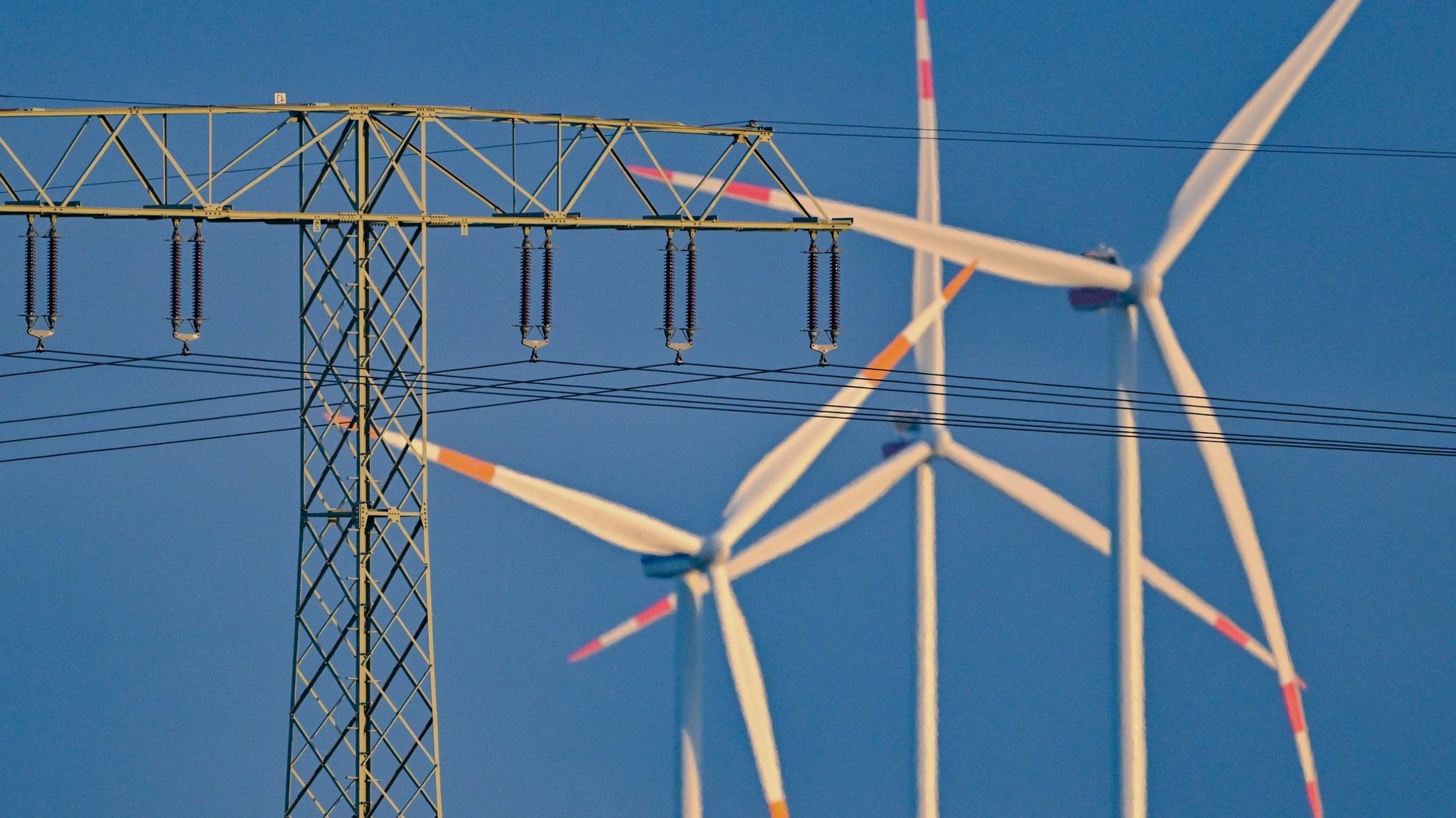 Ein Strommast mit einer Hochspannungsleitung steht vor Windenergieanlagen