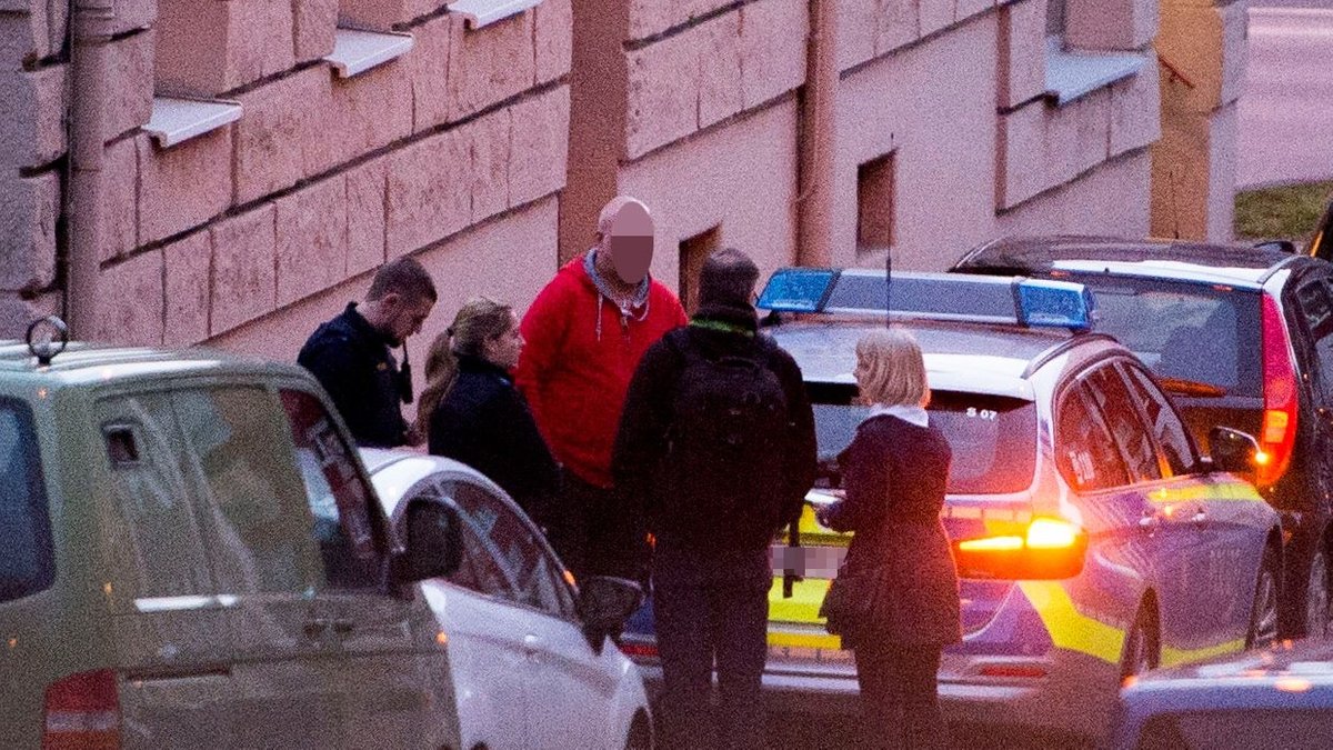 Eine Gruppe Menschen steht an einem Polizeiauto. Ein blonder Mann in roter Jacke ist gepixelt.