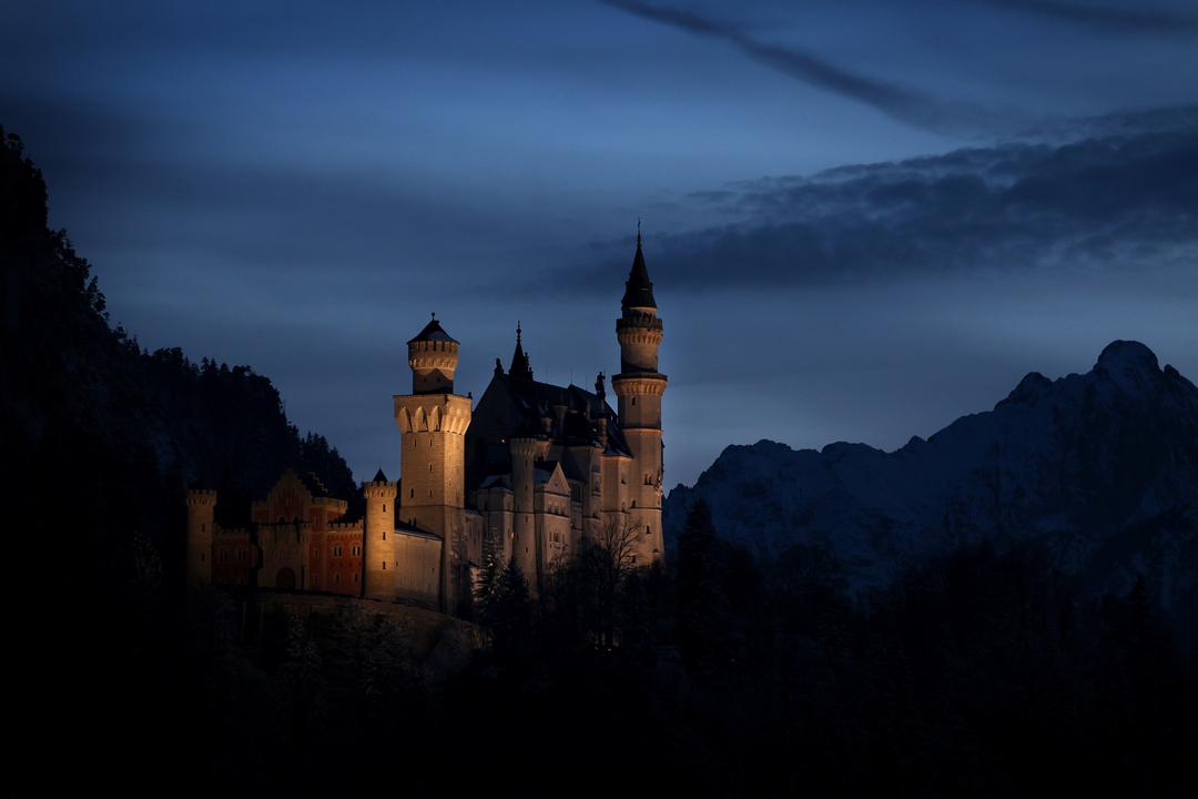 Bayern, Schwangau: Das Schloss Neuschwanstein steht nach Sonnenuntergang zur Blauen Stunde vor dem Panorama der Alpen.