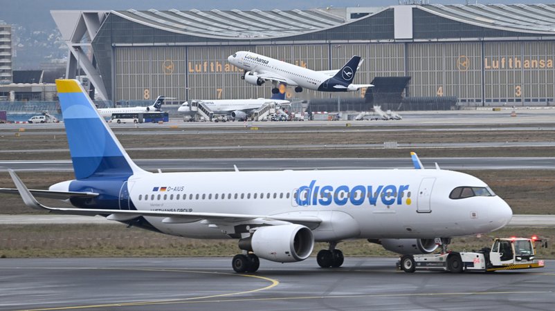 Während eine Maschine der Lufthansa startet, wird ein Passagierflugzeug der Discover Airlines am Frankfurter Flughafen von einem Schlepper gezogen. | Bild: dpa-Bildfunk/Arne Dedert Während eine Maschine der Lufthansa startet, wird ein Passagierflugzeug der Discover Airlines am Frankfurter Flughafen von einem Schlepper gezogen.