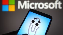 Ein Smartphone, auf dem Karl Klammer (der ehemalige Office-Assistent) zu sehen ist, im Hintergrund ein Logo von Microsoft | Bild:picture alliance / ZUMAPRESS.com | Pavlo Gonchar