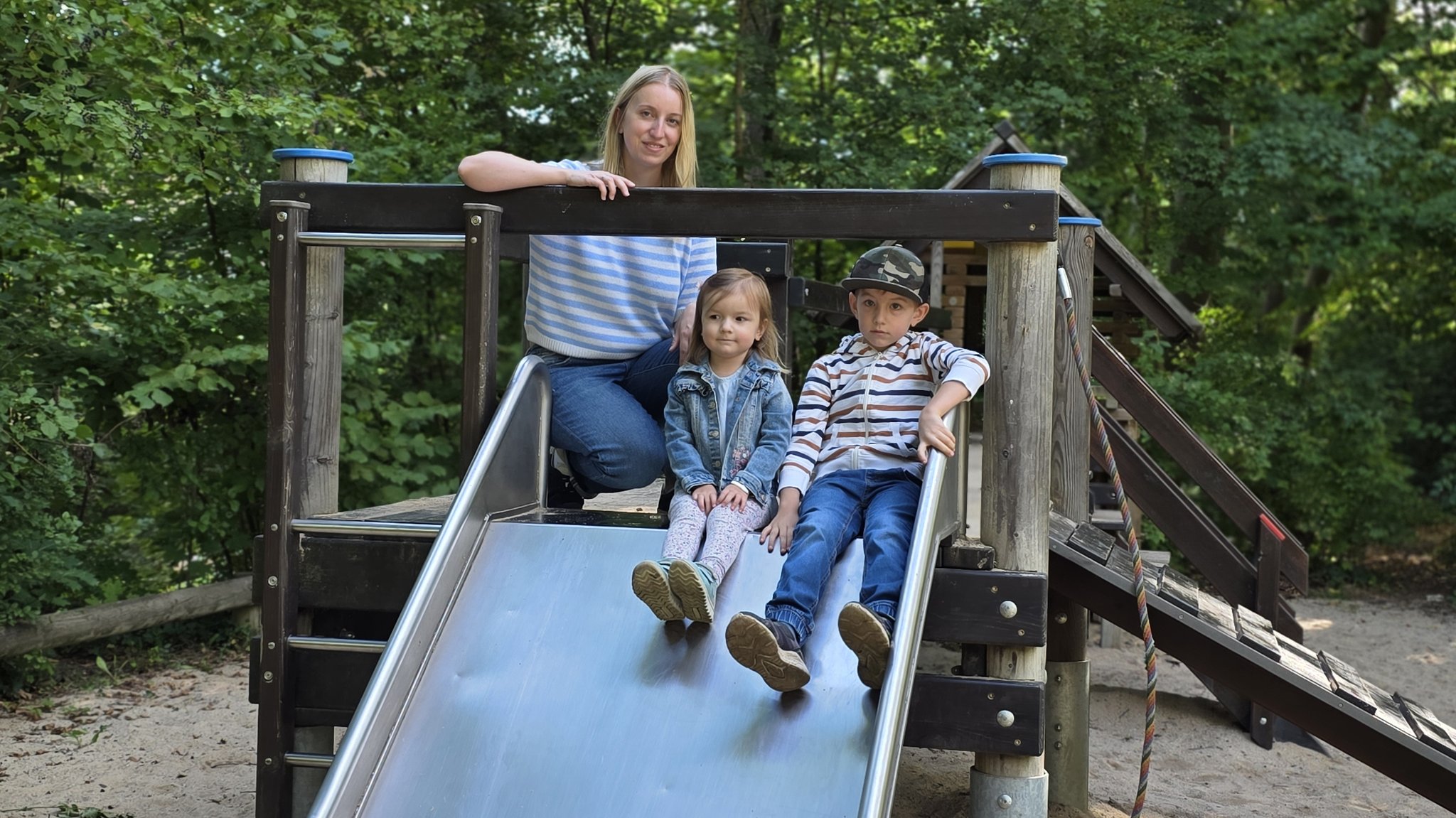 Lena sitzt neben ihrem Bruder und ihrer Mutter auf einer Rutsche auf dem Spielplatz.