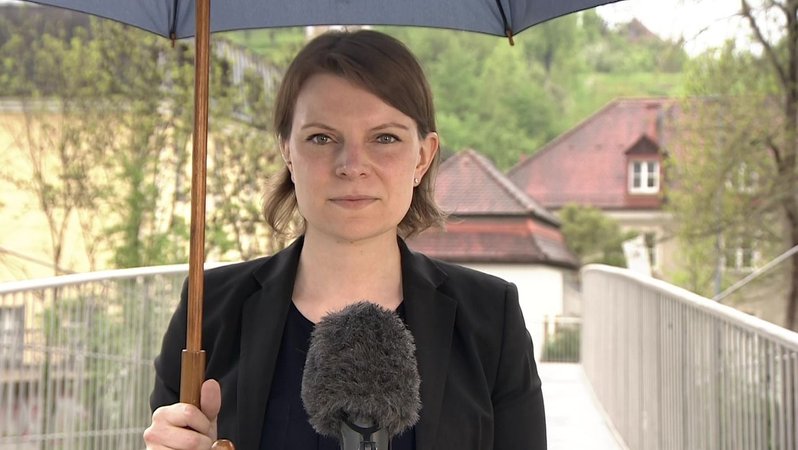 Emmi Zeulner steht mit Regenschirm auf einer Brücke vor einem Mikrofon.