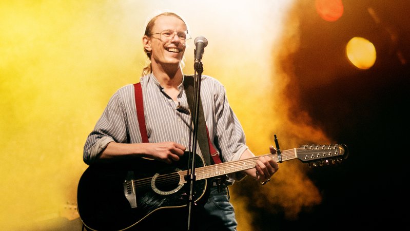 Mann mit Pferdeschwanz und Gitarre vor einem Mikrofon auf einer beleuchteten Bühne: Alexander Scheer als Liedermacher "Gundermann" 