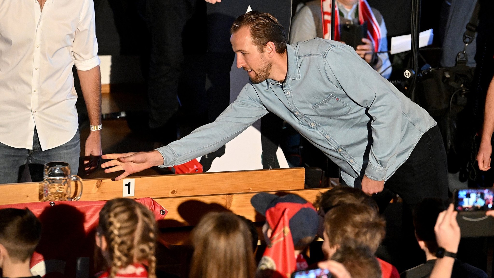 28.01.2024, Bayern, Kirchweidach: Fußball: Bundesliga, FC Bayern München. FC-Bayern-Spieler Harry Kane aus England schiebt einen Maßkrug beim Maßkrug-schieben im Fanclub «Die Roten» in Kirchweidach. Er erhielt dort eine Trophäe für seinen Sieg bei einer bayerischen Miniolympiade in Form einer Torjägerkanone, mit der er auch Schnupftabak genießen könnte. Die Stars des FC Bayern holten ihre wegen des Winterwetters im Dezember verschobenen Fanclubbesuche nach. 