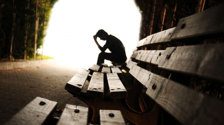 Ein Mensch sitzt zusammengesunken auf einer Bank. Im Vordergrund ist es düster, im Hintergrund scheint ein grelles Licht (Symbolbild) | Bild:stock.adobe.com/hikrcn