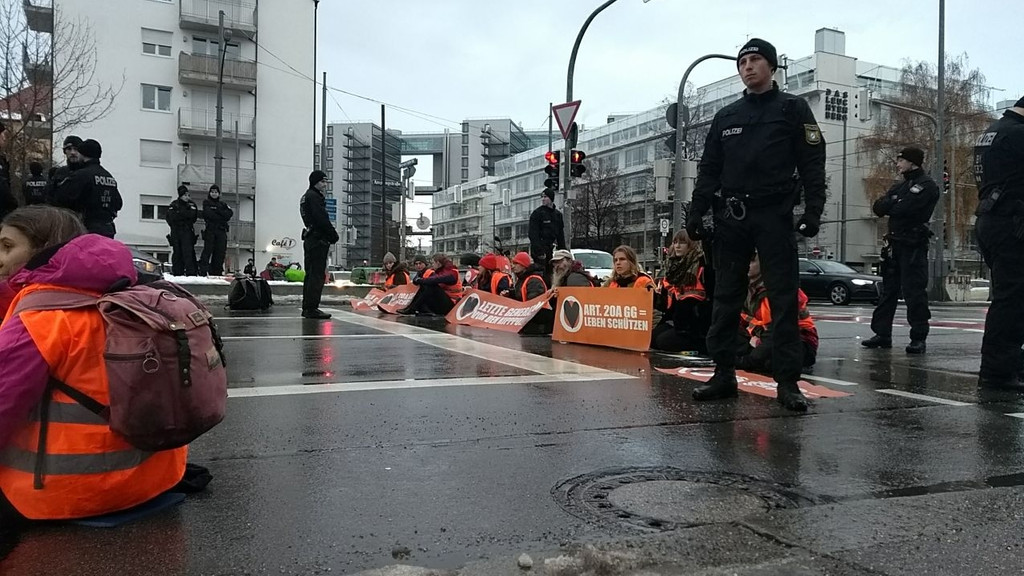 Zu sehen sind Demonstrierende der Gruppe "Letzte Generation" auf einer Straße nahe des Ostbahnhofs. Sie halten Transparente hoch, um sie herum stehen Polizisten.