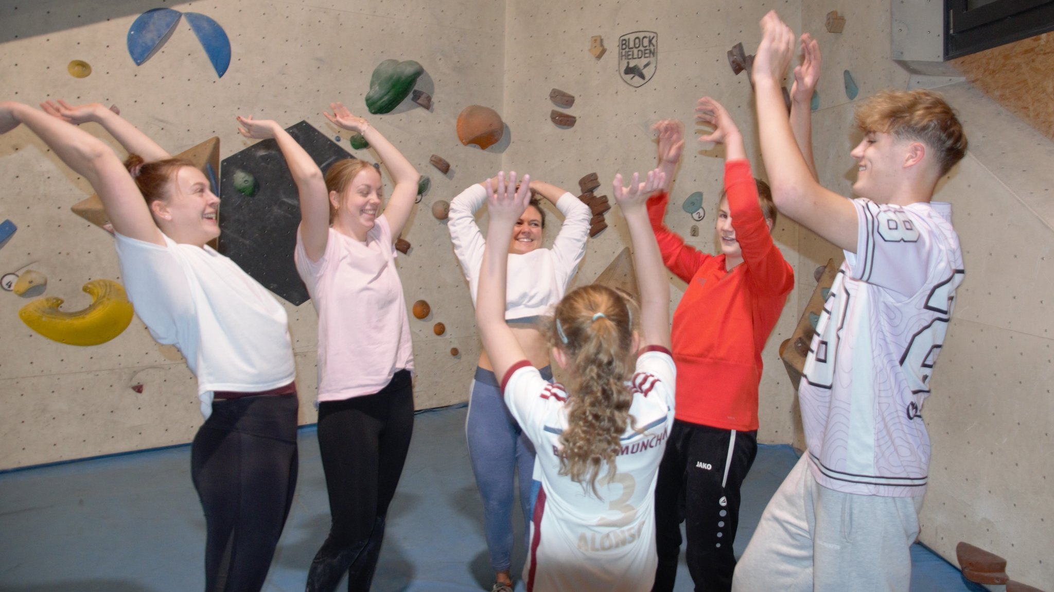 Sechs Jugendliche strecken ihre Arme in einer Boulderhalle freudig nach oben.