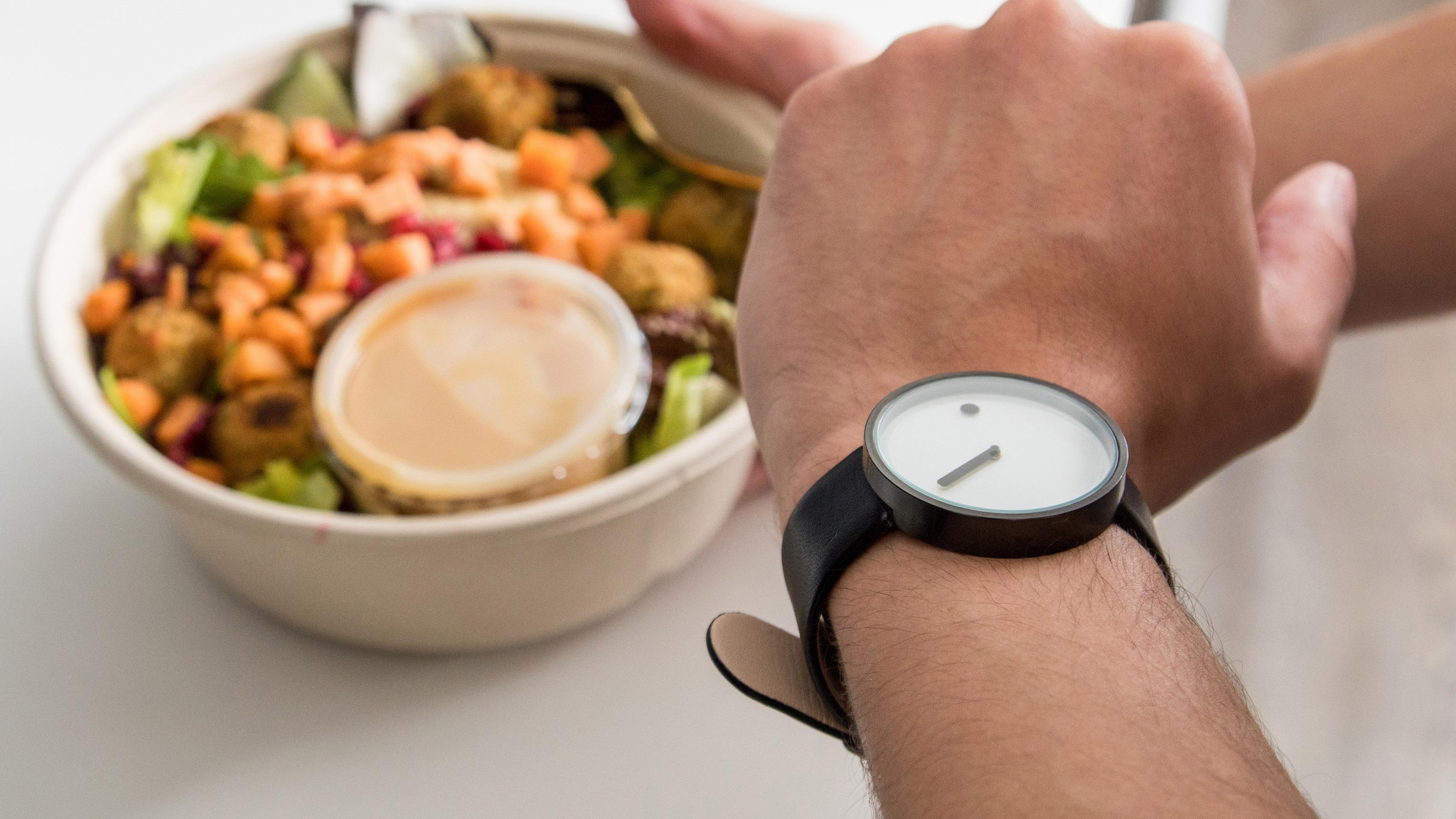  Ein junger Mann schaut am in der Küche auf seine Armbanduhr, während er nach einem Salat greift.