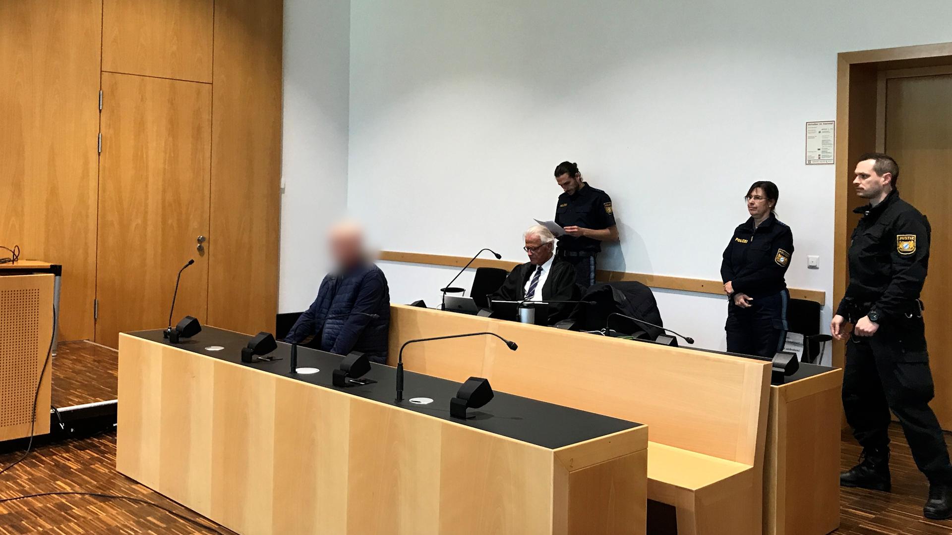 Der Angeklagte (links) im Gerichtssaal, im Hintergrund unter anderem Polizisten