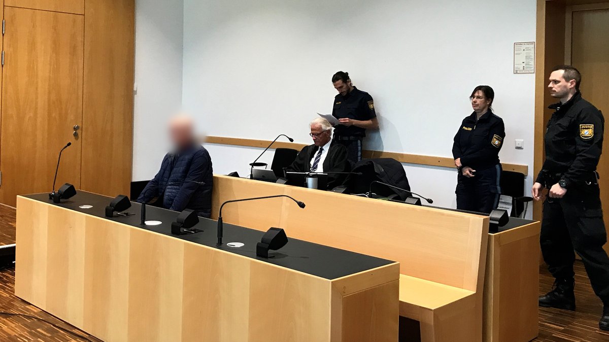 Der Angeklagte (links) im Gerichtssaal, im Hintergrund unter anderem Polizisten