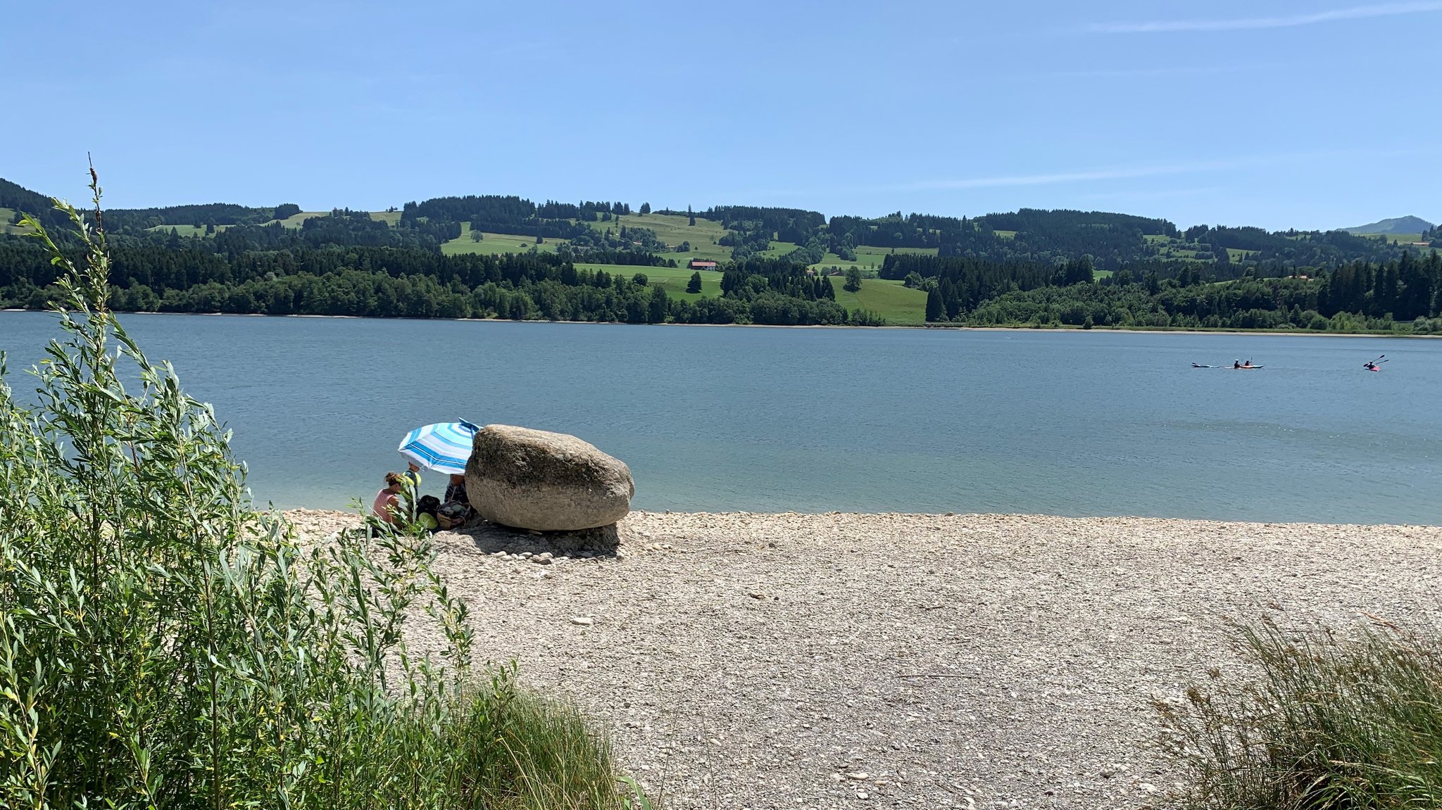 Blick auf den Rottachsee vom grünen Ufer aus - wegen des Niedrigwassers liegt zwischen grün und Wasser nun ein breiter Kiesstreifen. Darauf sitzen Badegäste unter einem gestreiften Sonnenschirm.