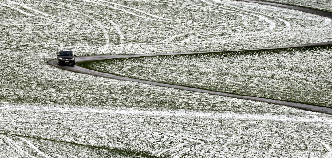 Ein Auto fährt im Allgäuer Voralpenland zwischen den teilweise mit Schnee bedeckten Wiesen.