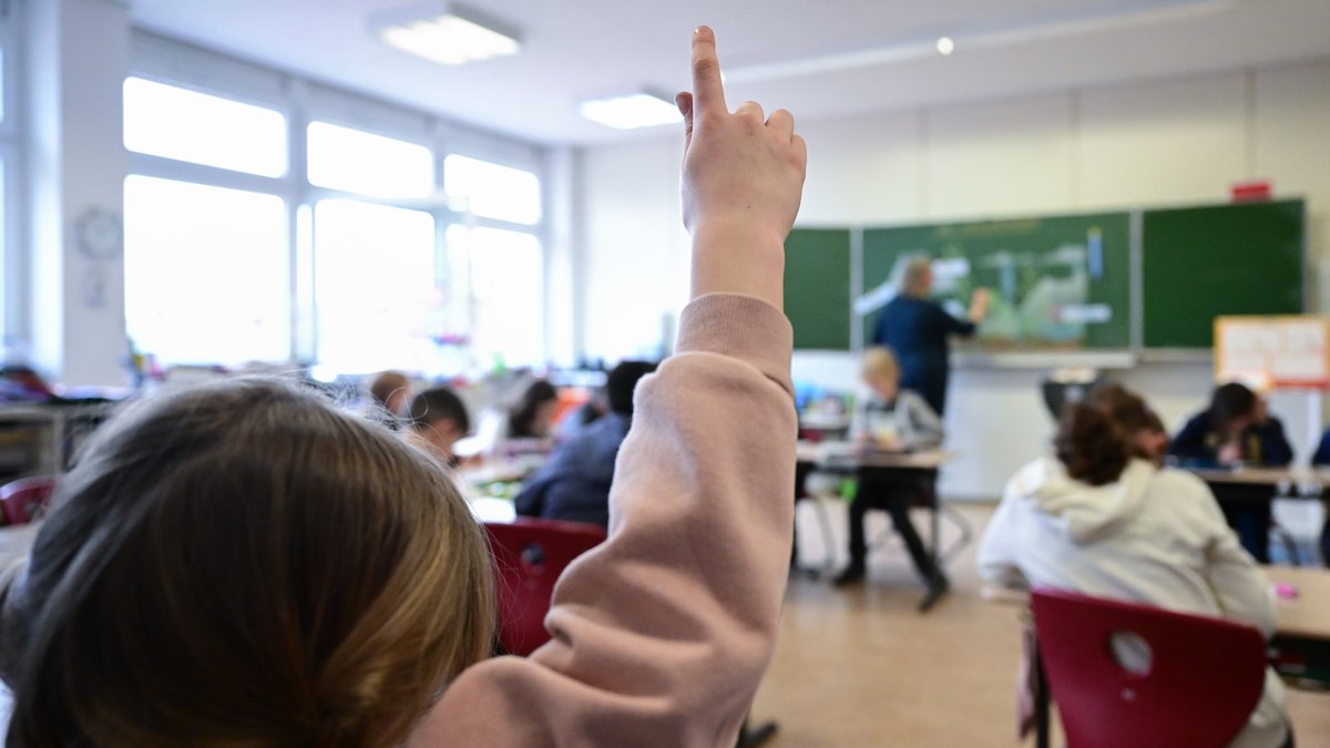 Ein Kind hebt den Finger im Unterricht an einer Grundschule. Im Hintergrund sieht man eine Lehrerin an der Tafel stehen.