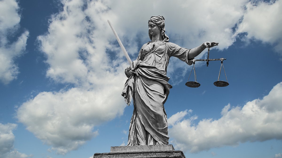Statue einer Justizia mit Waage und Schwert vor einem Wolkenhimmel.