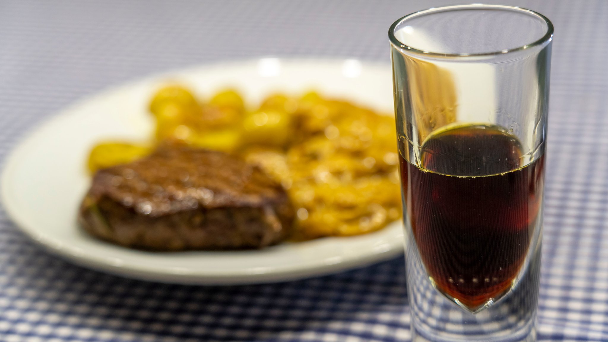 Ein Gläschen Kräuterschnaps im Vordergrund - im Hintergrund ein Teller mit Fleisch und Bratkartoffeln