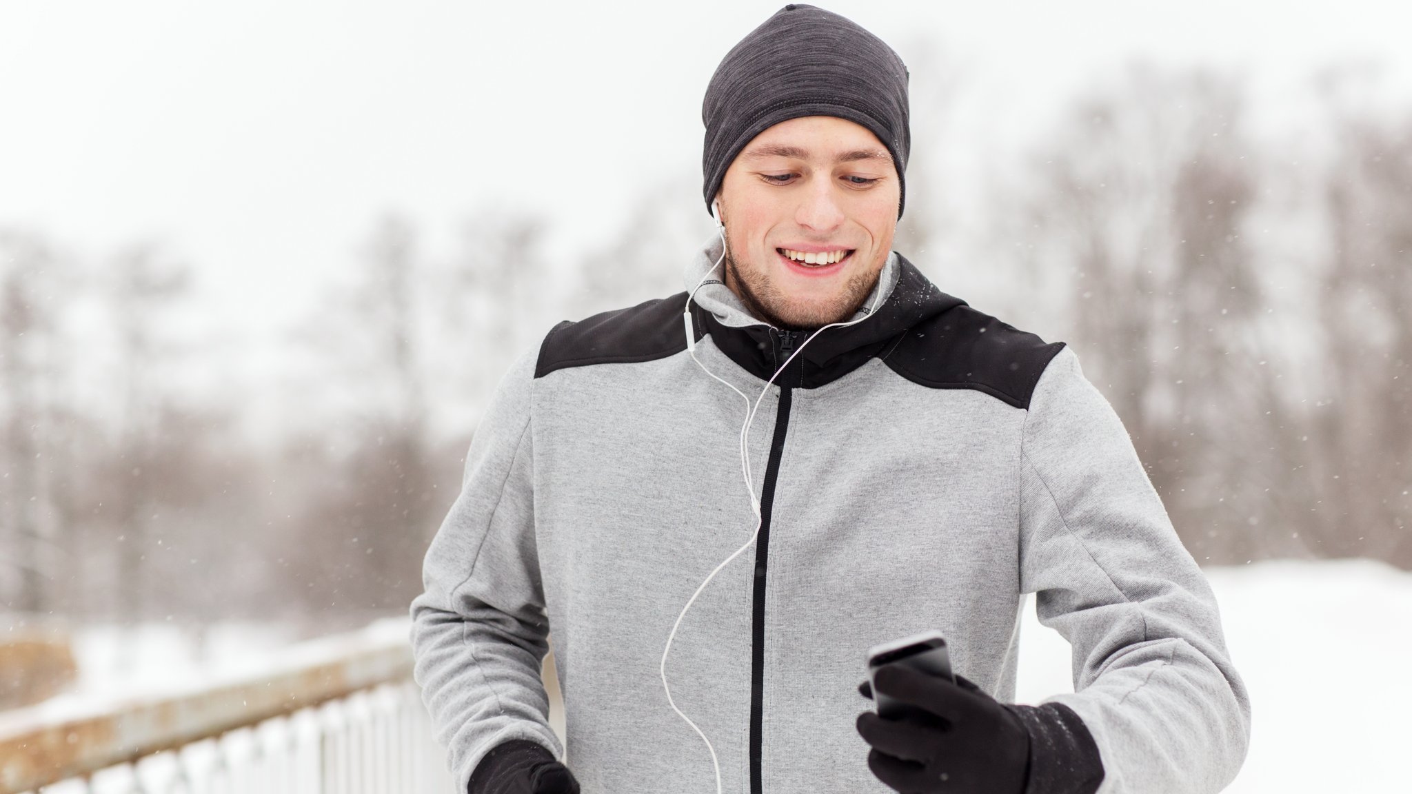 Mann in Frontalansicht joggt im Schnee mit Mütze, Handschuhen und Kopfhörern im Ohr