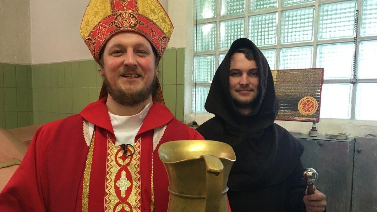 Ein Mann, gekleidet wie ein Bischoff, hält einen goldenen Krug in der Hand. Hinter ihm steht ein Mann mit einer Kapuze über dem Kopf und einem dunklen Umhang, ein "Aprostel". 