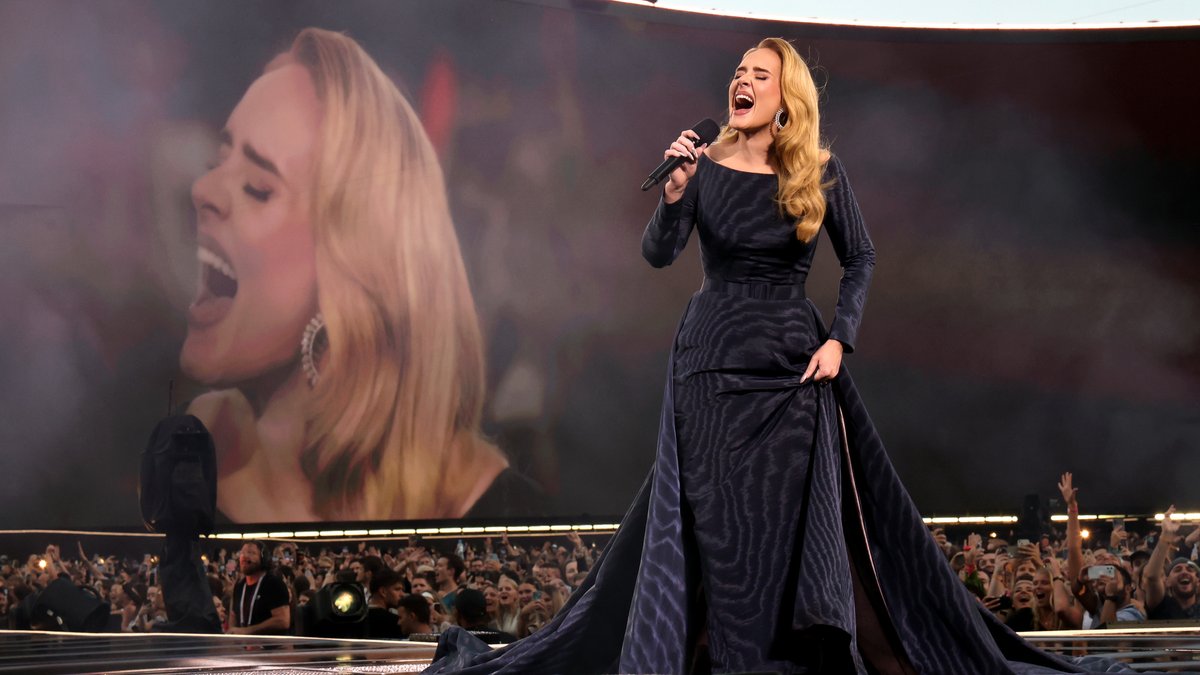 Popstar Adele in dunklem Kleid, im Hintergrund ihr Gesicht auf großer Videoleinwand