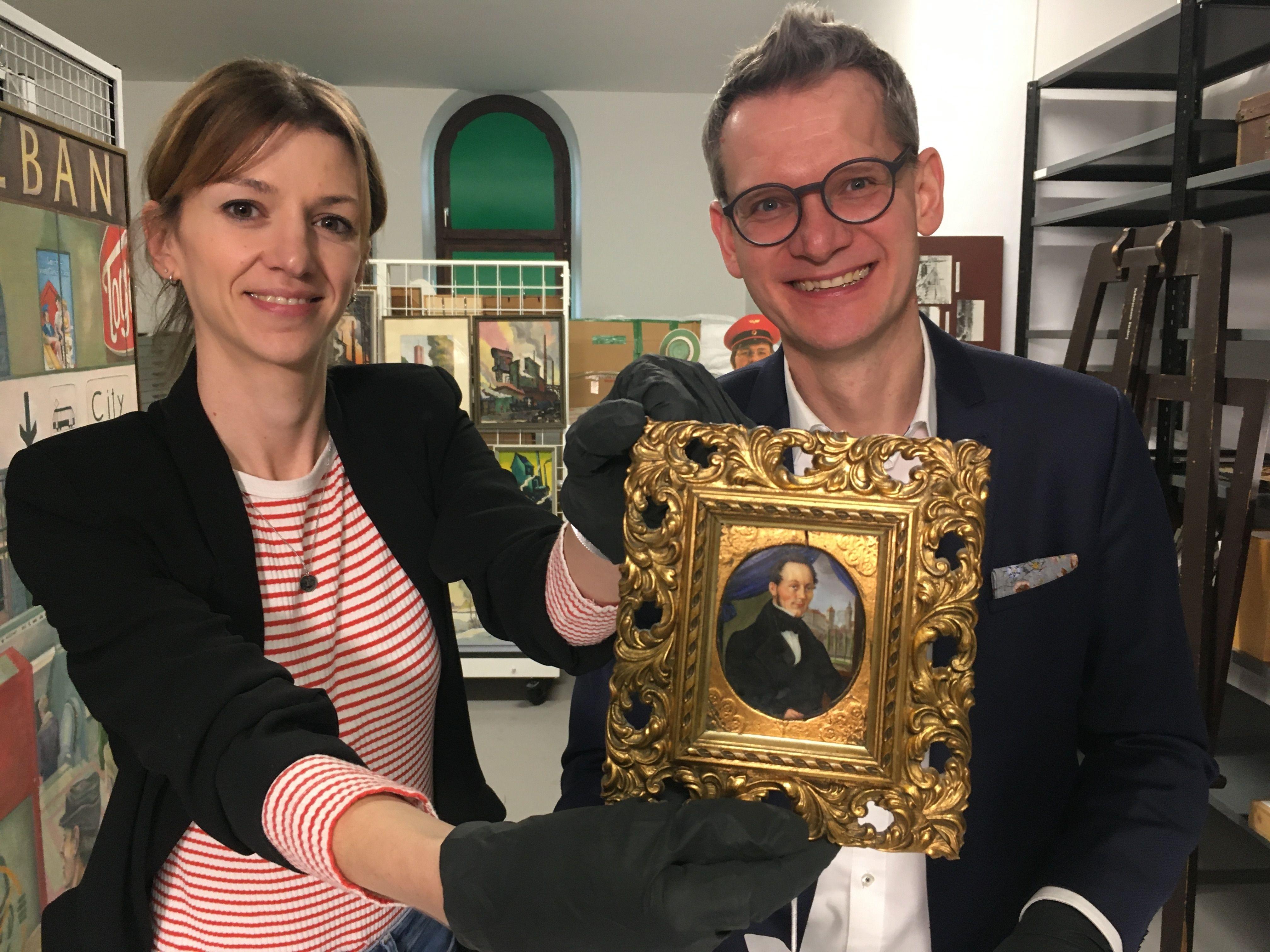 Zwei Museumsmitarbeiter halten ein historisches Portrait im Goldrahmen hoch.